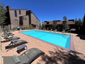 Una piscina rectangular con superficie de agua azul cristalina, rodeada por un patio de ladrillo con sillones de descanso y una pequeña mesa.