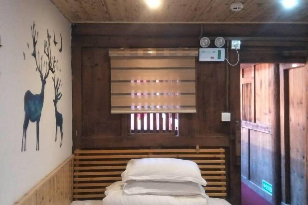 A wooden cabin room with a bed, a wall mural of deer, a window with blinds, and a wall-mounted thermostat.