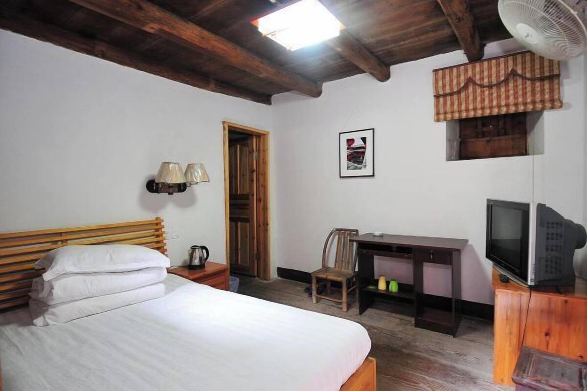 A bedroom with a bed, wooden headboard, bedside table with a lamp, a desk with a chair, a television, and a small window with a curtain.