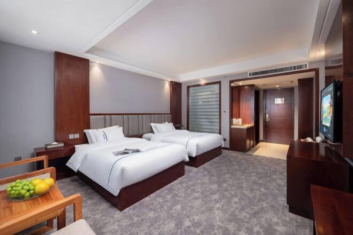 A hotel room with two beds, a desk, a TV, and a small table with fruit.