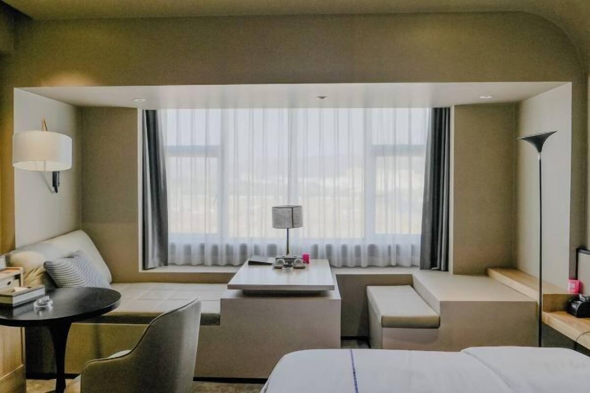 A hotel room with a bed, a desk, a chair, a sofa, a lamp, and a large window with curtains.