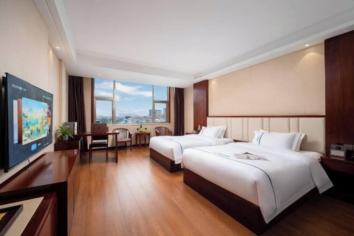 A hotel room with two beds, a desk, a chair, a TV, and a large window with a city view.