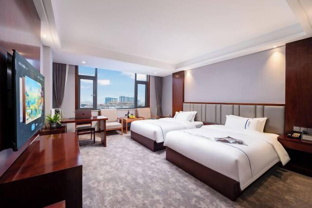 A hotel room with two beds, a desk, a TV, and a large window with a city view.