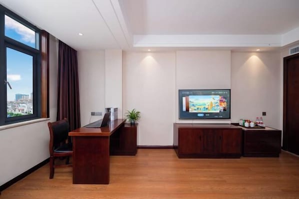 A modern hotel room with a wooden desk, a flat-screen TV mounted on the wall, and a large window offering a city view. - Mengzi Yufeng Hotel (Mengzi)