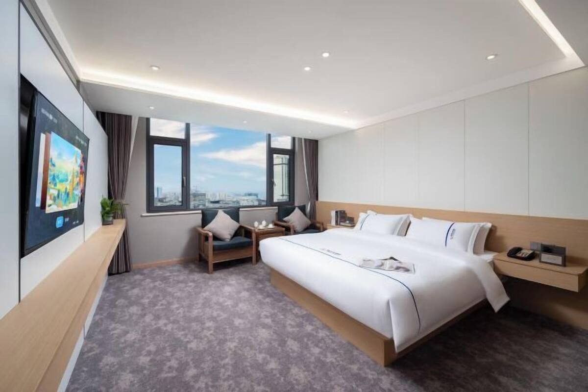 A modern hotel room with a large bed, a desk, a chair, a view of the city, and a large window.