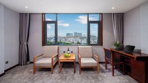 Une chambre moderne avec vue sur la ville, deux canapés, une petite table avec une bouteille et des fruits, et un bureau avec un ordinateur portable.