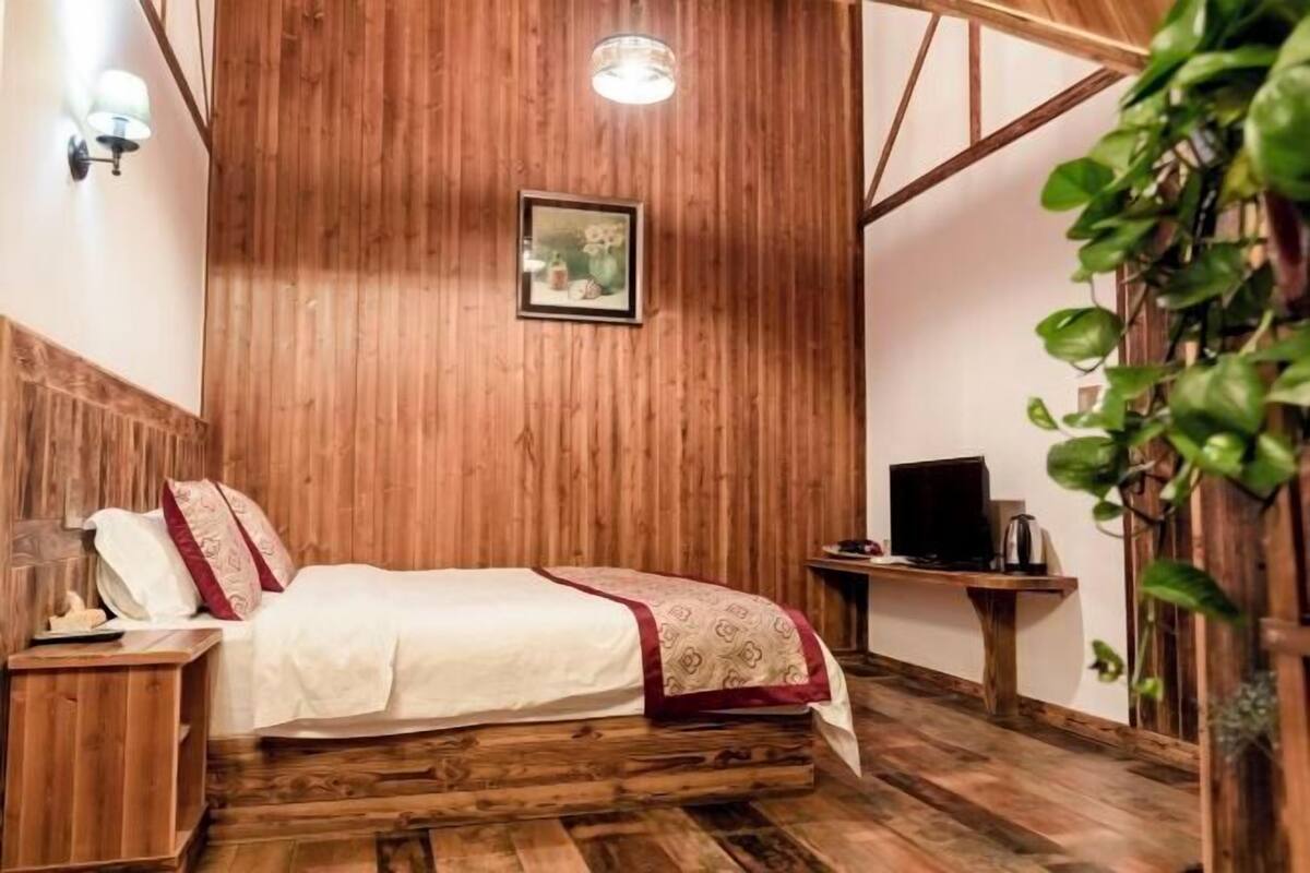 Una habitación con paneles de madera, una cama, una mesita pequeña, un televisor y una lámpara fijada a la pared.