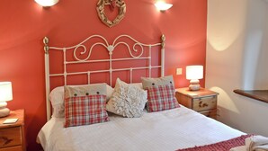 A bed with white bedding, a red and white checkered pillow, and a white floral pillow. There are two bedside tables with lamps, a wooden nightstand, and a wall-mounted light fixture.