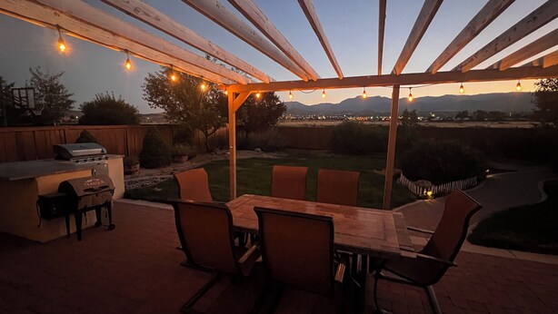 An outdoor patio with a wooden pergola, string lights, a grill, and a view of the landscape.