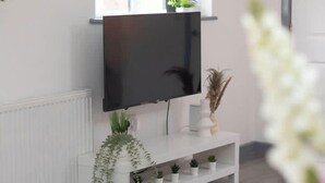Un salon moderne avec un téléviseur à écran plat fixé sur un meuble blanc, une fenêtre au-dessus et une table à manger avec de l’assiette blanche et un vase de fleurs.
