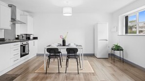 Une cuisine moderne avec des meubles blancs, une table à manger avec deux chaises, un réfrigérateur et une fenêtre donnant sur l’extérieur.