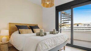 A bedroom with a large bed, a nightstand with a lamp, and a view of a building through a large window. - Casa Carmen - Rooftop Luxury Penthouse Estepona (Estepona)