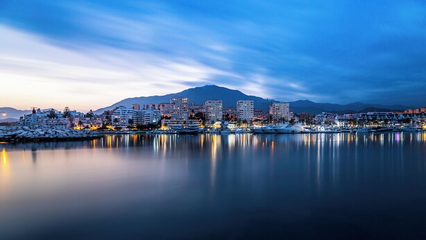 A coastal cityscape at dusk with a marina, buildings, and a mountain in the background. - Casa Carmen - Rooftop Luxury Penthouse Estepona (Estepona)