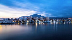 Un paisaje urbano costero al anochecer con un puerto deportivo, edificios y una montaña al fondo.