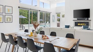 A modern dining area with a wooden table, black chairs, and a sofa. There is a TV mounted on the wall and a fireplace. - The Polo Villas by AvantStay (La Quinta)