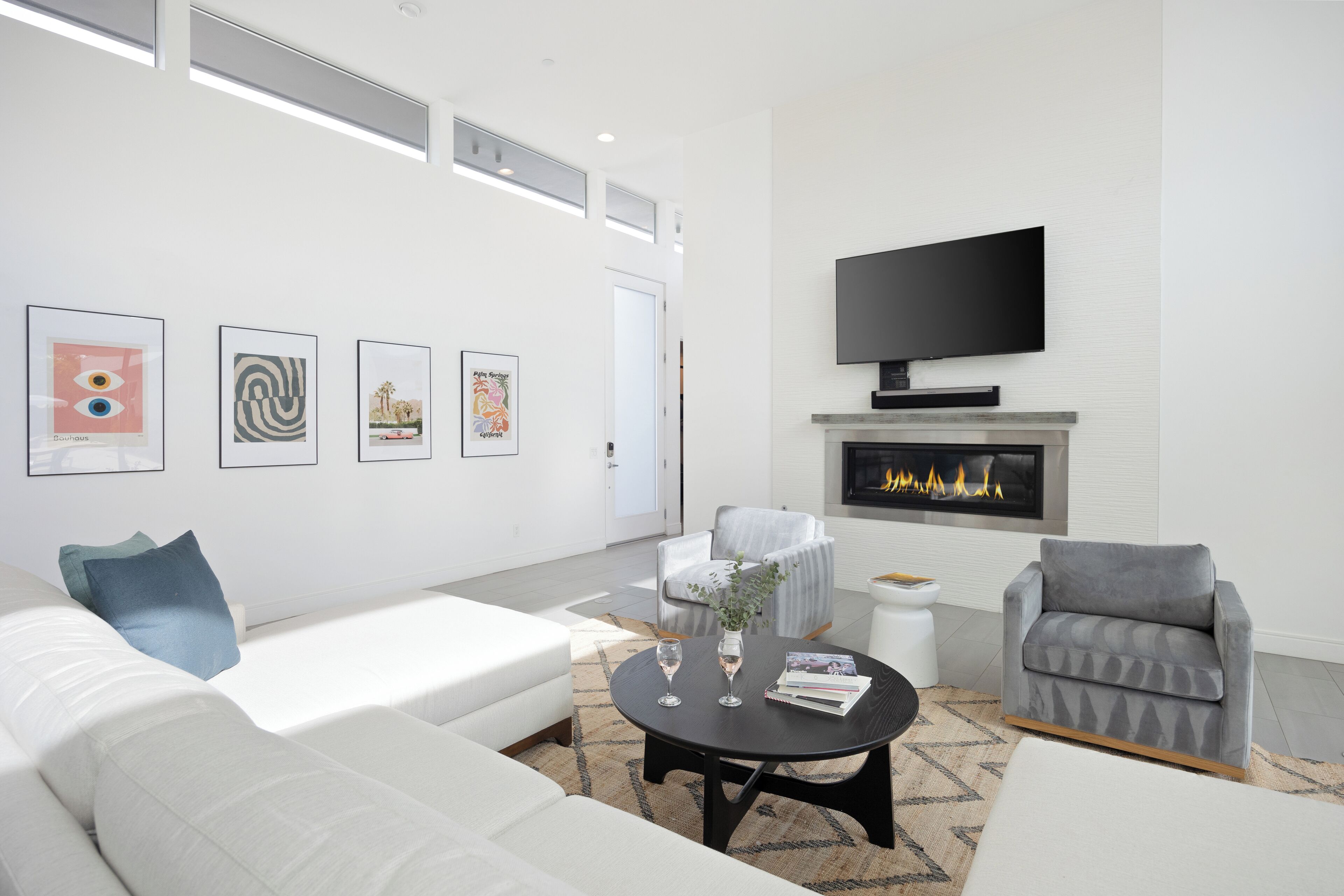 A modern living room with a white sofa, grey armchair, and a black coffee table. There are framed artworks on the wall, a flat-screen TV above a fireplace, and a small side table with a vase and books.