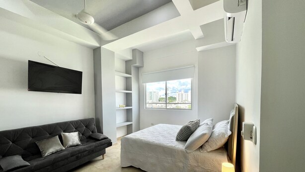 Un dormitorio moderno con una cama, un sofá oscuro, un televisor y una ventana con vistas a la ciudad.