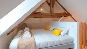 Ein Schlafzimmer mit schrägen Wänden, Holzbalken, einem Bett mit weißer Bettwäsche und gelben Kissen sowie einem zusammengerollten Handtuch auf dem Bett.
