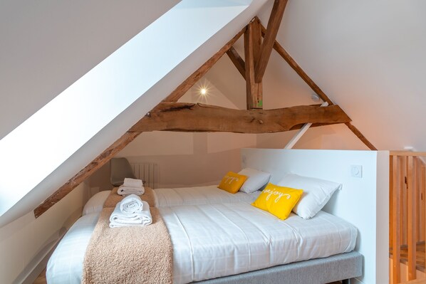 A bedroom with a sloped ceiling, wooden beams, a bed with white bedding and yellow pillows, and a rolled towel on the bed.