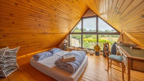 A cozy attic bedroom with a sloped wooden ceiling, a large window, a bed with pillows, a desk, and a piano. - La Renardière - Maison avec vue mer (Barneville-Carteret)