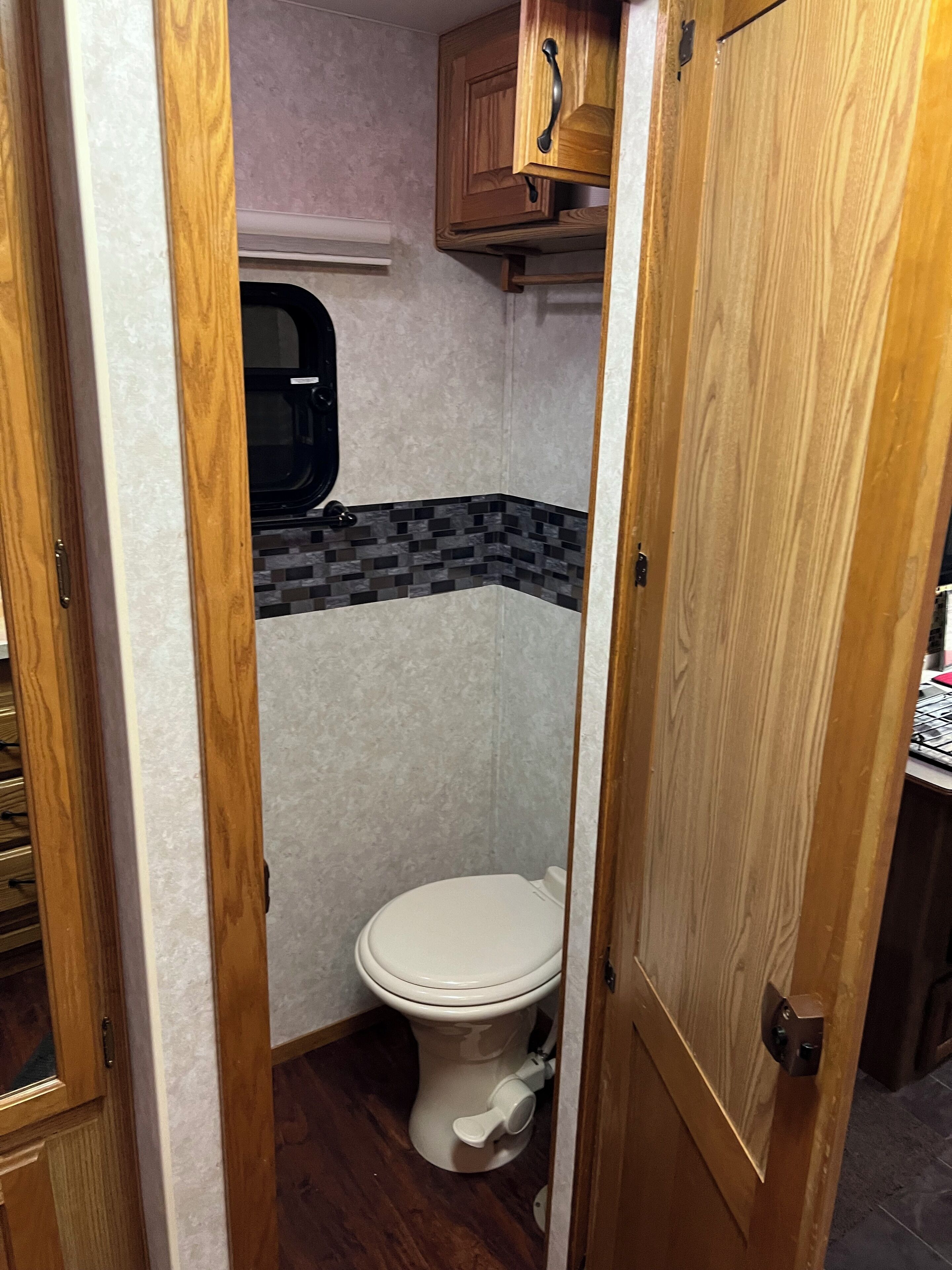 Een compacte badkamer met een wit toilet, een klein raam en een houten kast erboven.