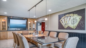 A modern dining area with a large glass table, comfortable chairs, a flat-screen TV, and a wall art piece of playing cards.
