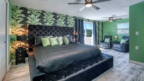 A bedroom with a large bed, a ceiling fan, a nightstand, and a view of the outside.