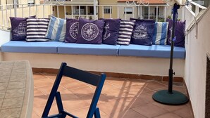 A terrace with a blue chair, a cushioned bench with patterned cushions, and a floor lamp.