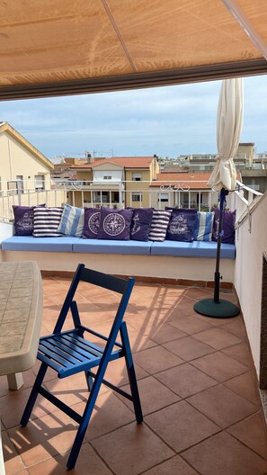 A terrace with a blue chair, a cushioned bench with patterned cushions, and a floor lamp.