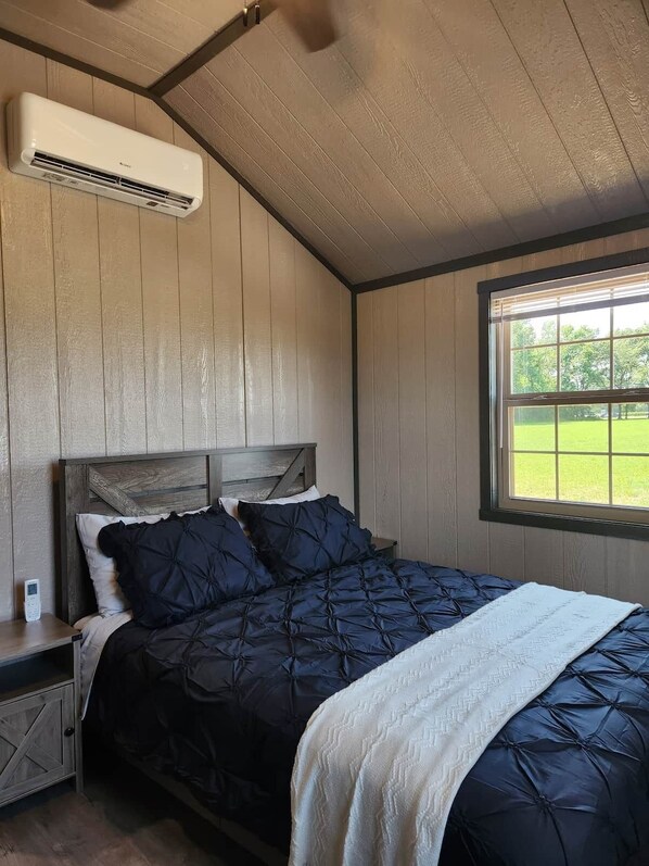 A bedroom with a bed, wooden walls, a window with a view, and an air conditioning unit.