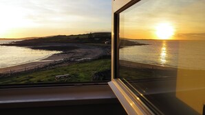 Vista desde una ventana a un paisaje costero con atardecer, zonas de hierba y una costa rocosa.