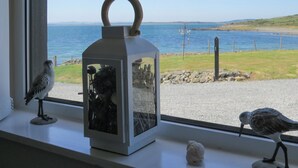 Una ventana con vista al mar, un farol blanco y dos figuras de pájaros en el alféizar.
