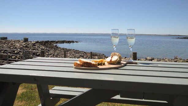 Una mesa de picnic con comida y dos vasos de vino blanco, con vista al mar tranquilo y a la costa rocosa.