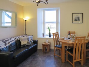 Sala de estar com sofá de couro preto, mesa de jantar com cadeiras, abajur e janela com vista.