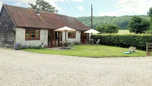A rustic house with a gravel driveway, a patio with a table and chairs, and a view of a green field and trees in the background. - The Dairy at Copper Beeches (Petersfield)