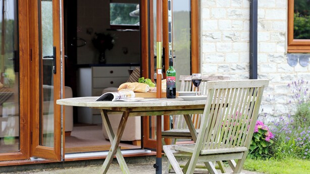 A wooden table and chair set under an umbrella, with a bottle and glass on the table. - The Dairy at Copper Beeches (Petersfield)