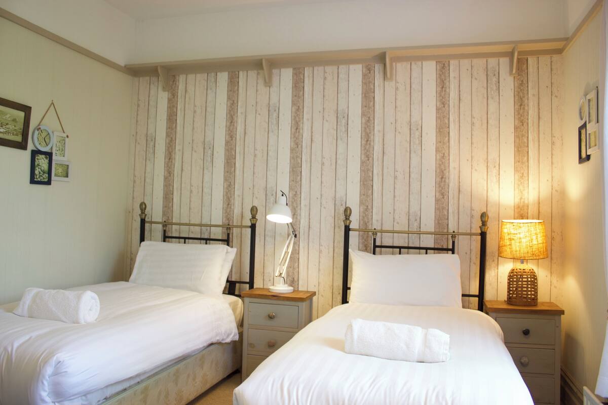 A room with two beds, each with a bedside table and lamp. The walls have a wooden panel design.