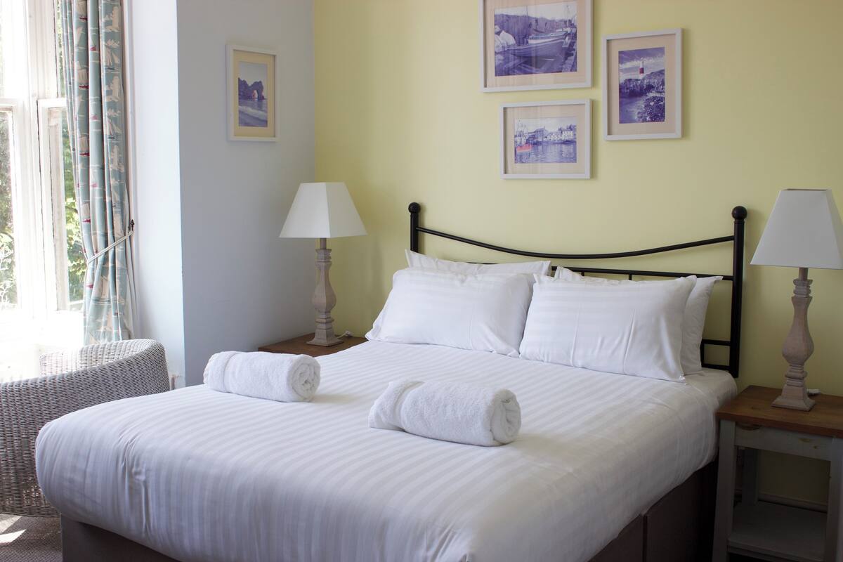 A neatly made bed with white linens and two rolled towels, a bedside table with a lamp, and a window with curtains.