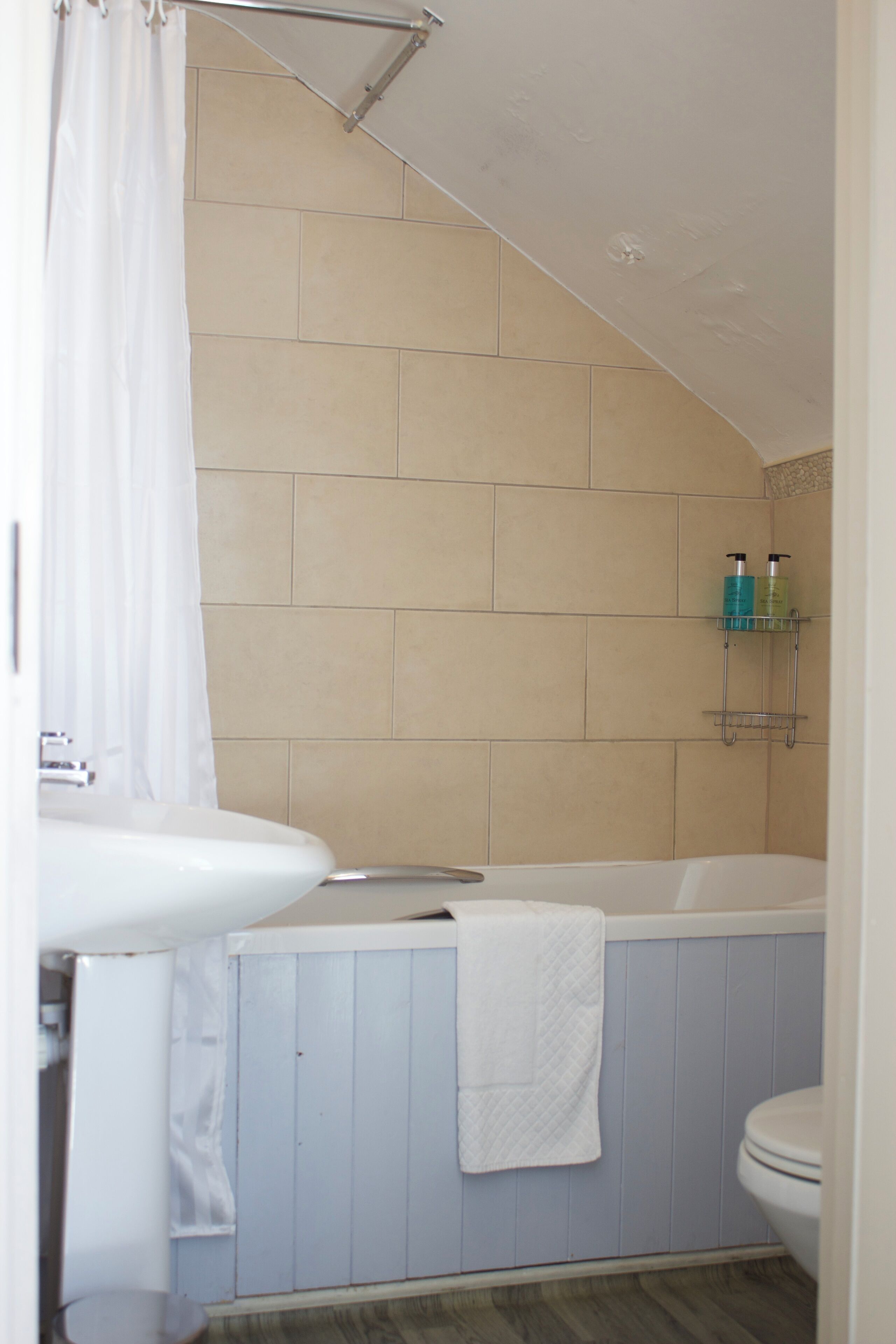 A bathroom with a bathtub, a sink, and a toilet. There is a shower curtain, a towel hanging on the wall, and a shelf with toiletries.