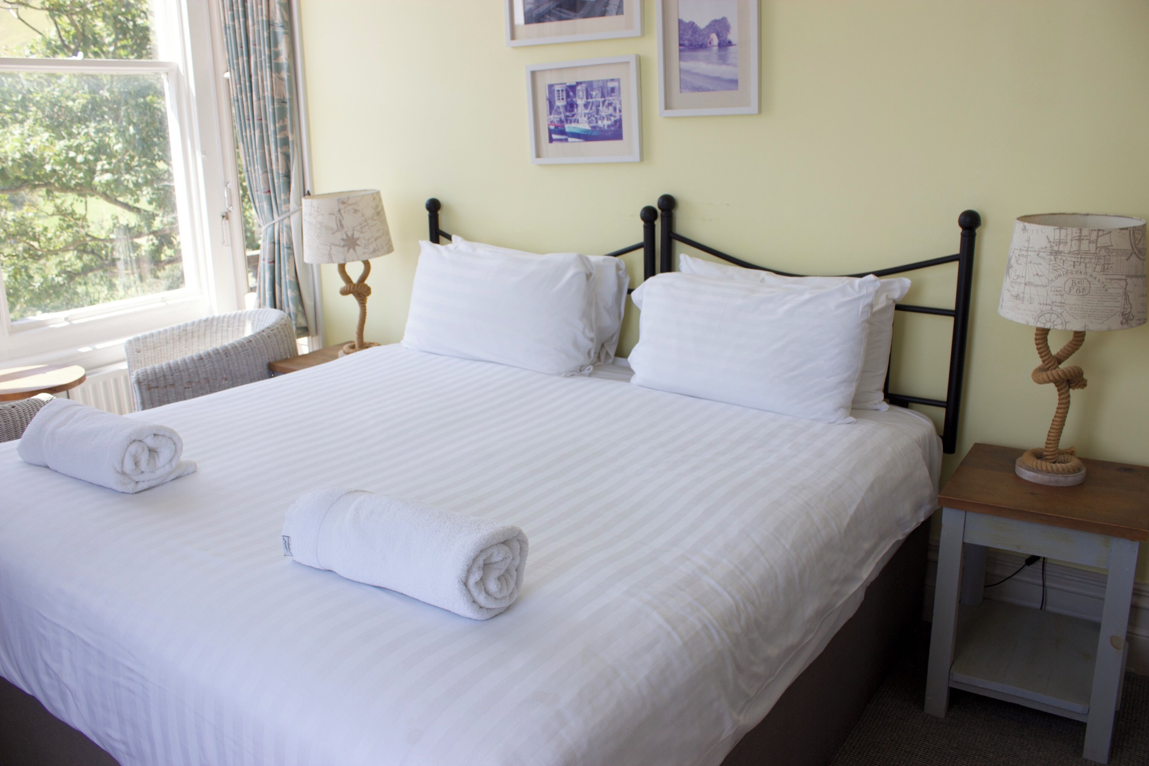 A neatly made bed with white linens and two rolled towels, a bedside table with a lamp, and a window with curtains.