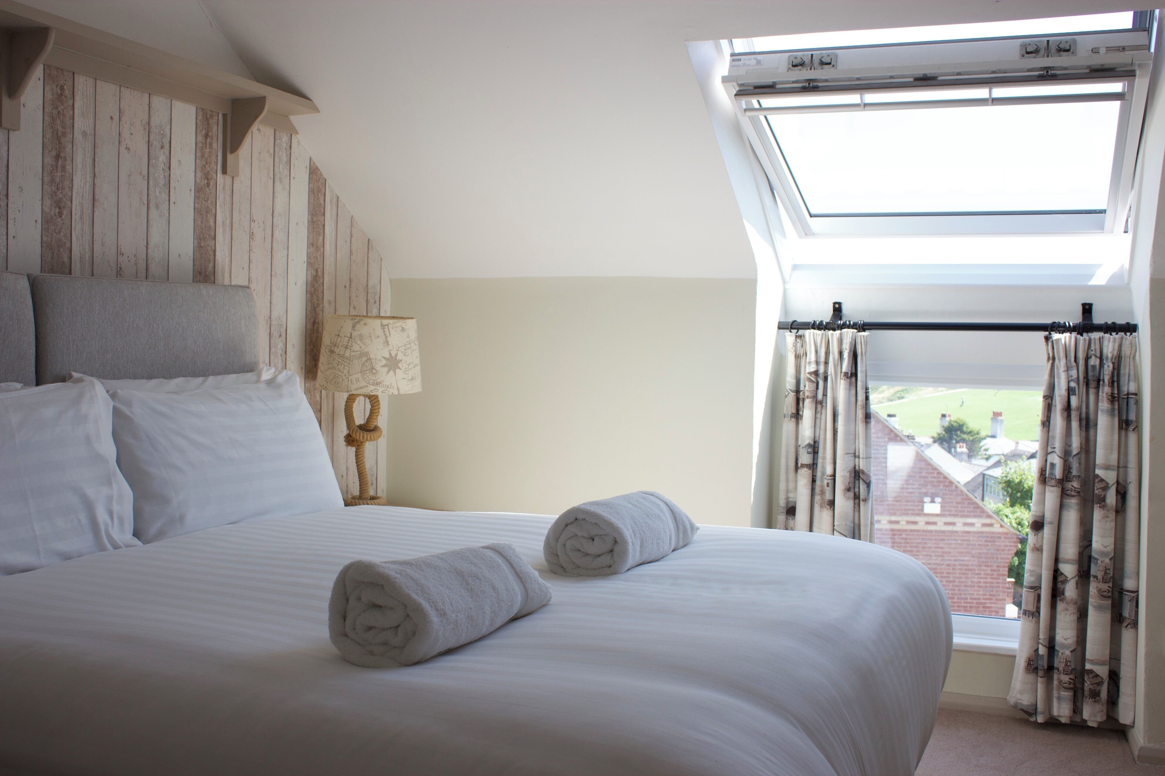 Ein Schlafzimmer mit einem großen Bett, zwei gerollten Handtüchern, einem Nachttisch mit Lampe und Blick auf ein Backsteingebäude durch ein Fenster mit Vorhängen.