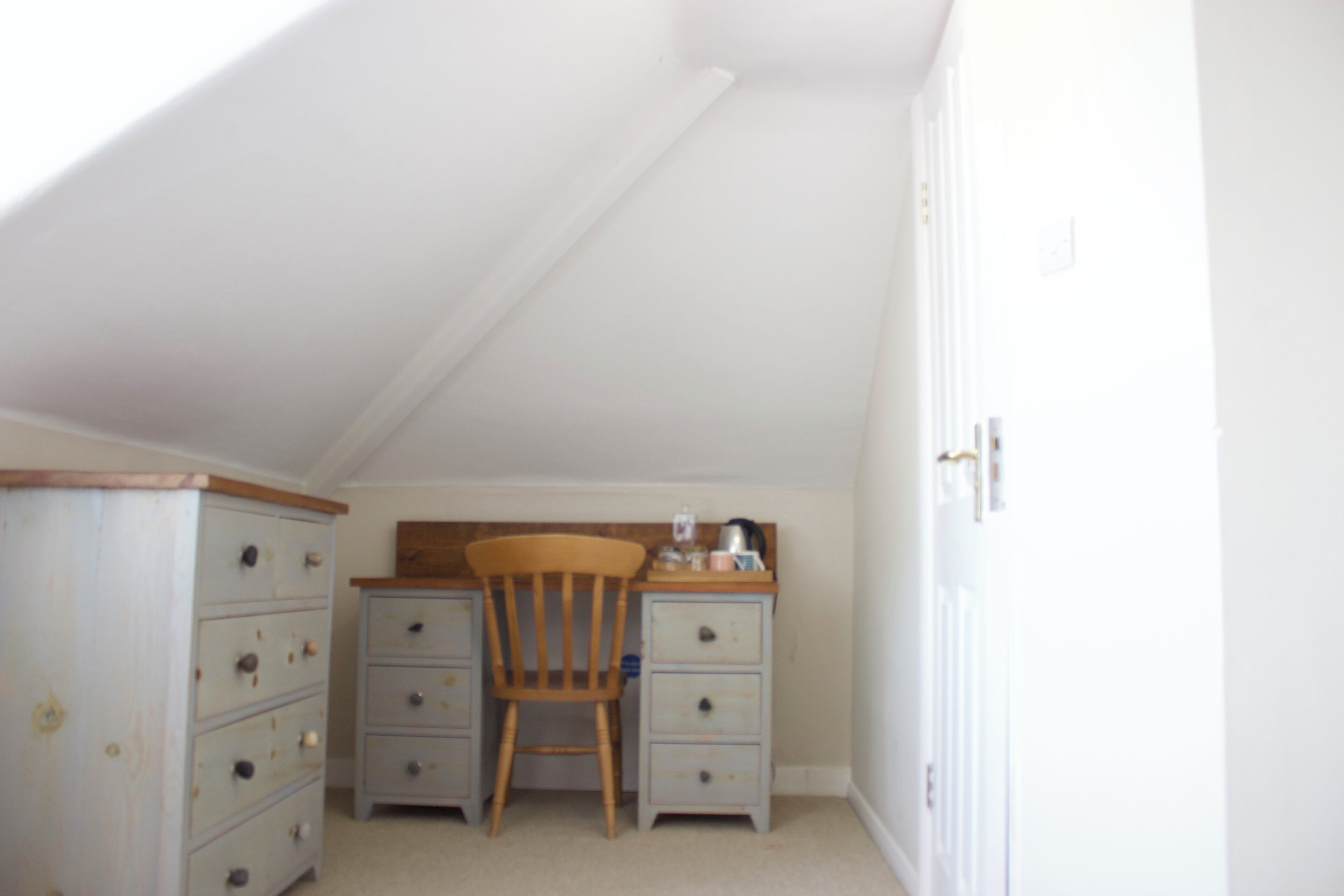 A small, well-lit room with a sloped ceiling, a wooden chair, a desk with drawers, and a dresser.