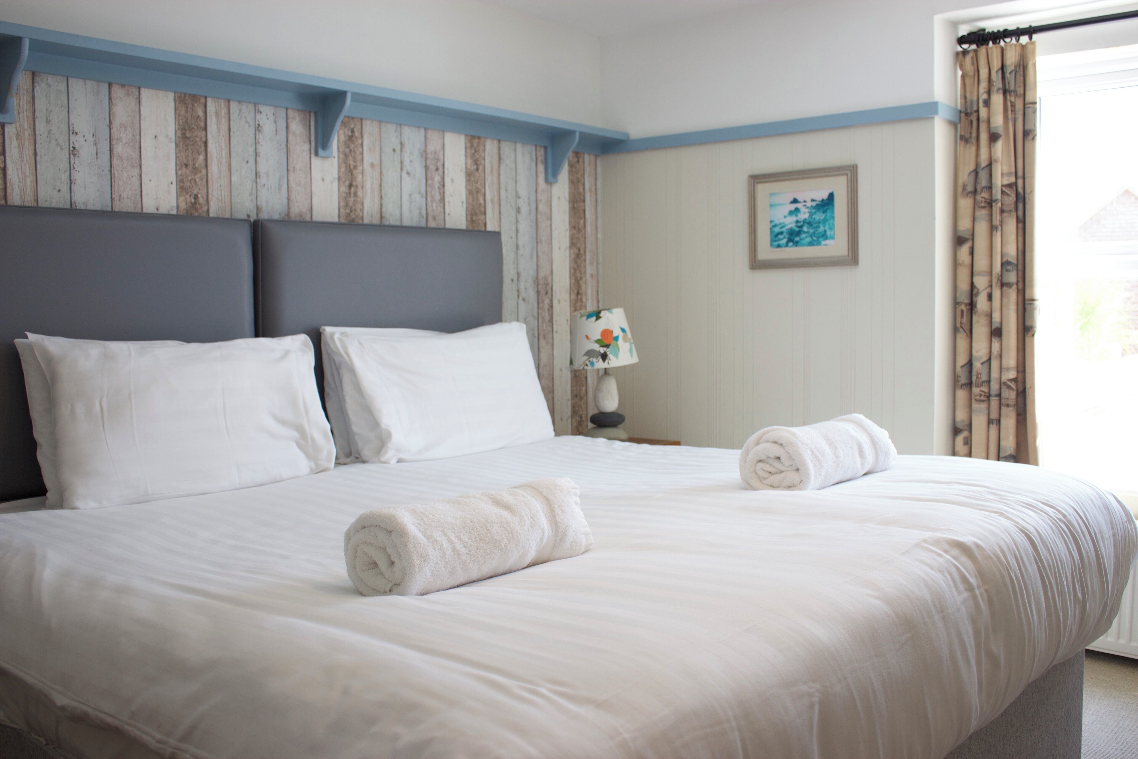 Een net opgemaakt bed met witte lakens en twee opgerolde handdoeken, een houten hoofdbord, een bedlampje en een ingelijst foto op de muur.