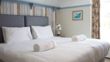 Een net opgemaakt bed met witte lakens en twee opgerolde handdoeken, een houten hoofdbord, een bedlampje en een ingelijst foto op de muur.