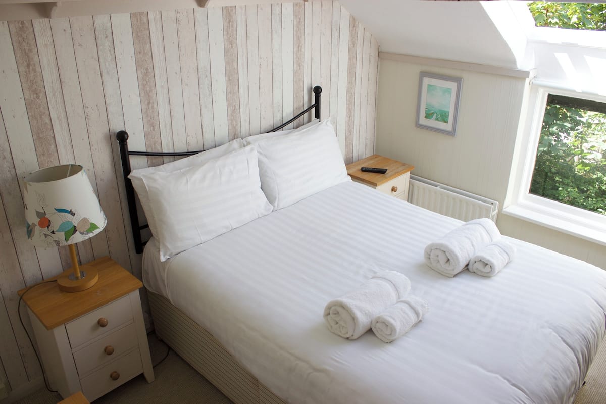 A neatly made bed with white linens and rolled towels, a bedside lamp, and a small wooden nightstand with drawers.