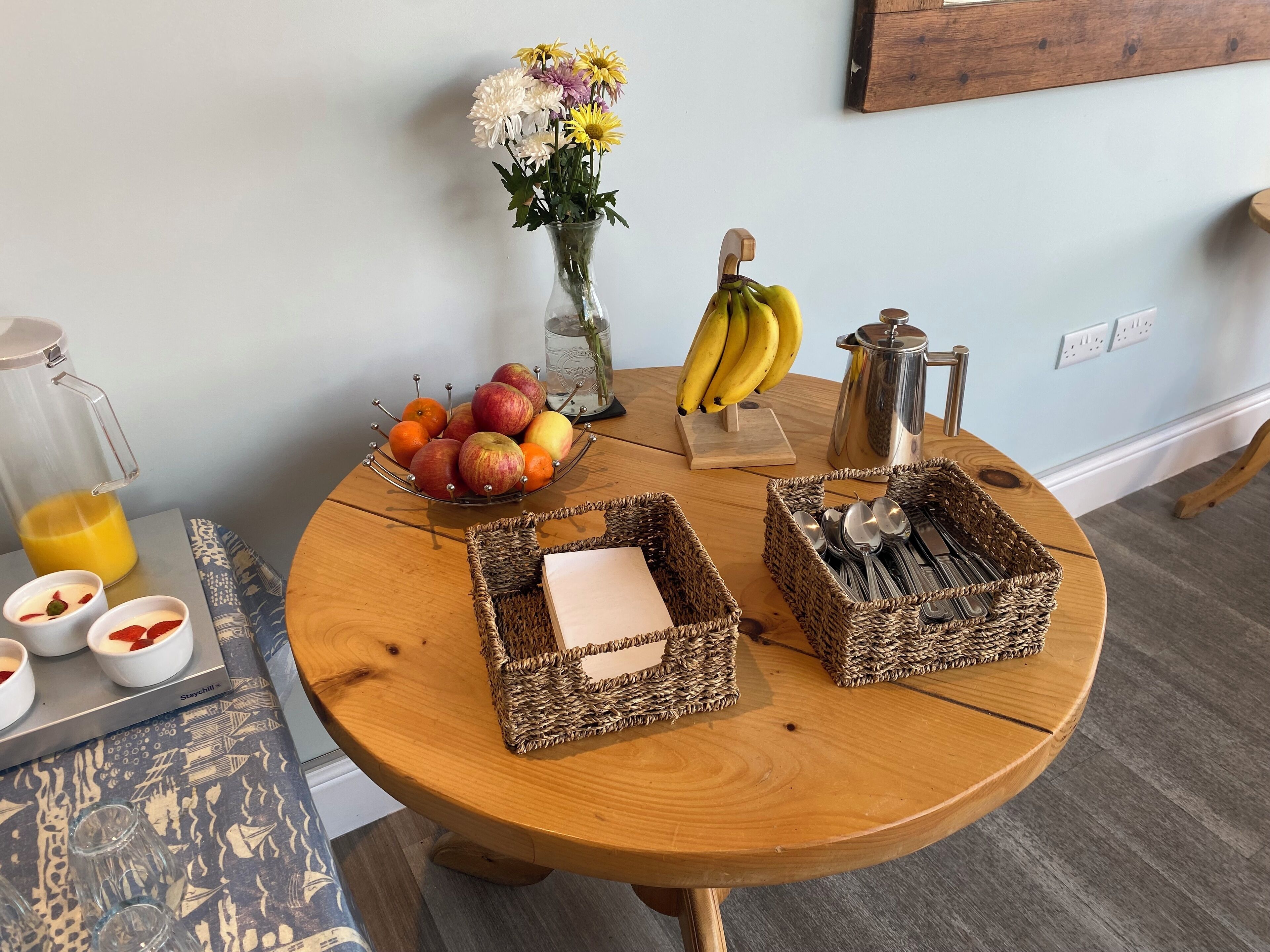 A wooden table with a fruit bowl, a vase with flowers, a coffee pot, and two woven baskets containing utensils.
