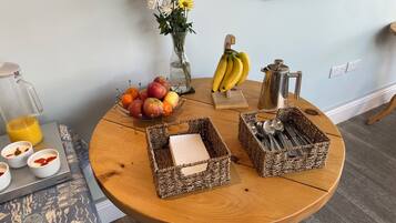 A wooden table with a fruit bowl, a vase with flowers, a coffee pot, and two woven baskets containing utensils.