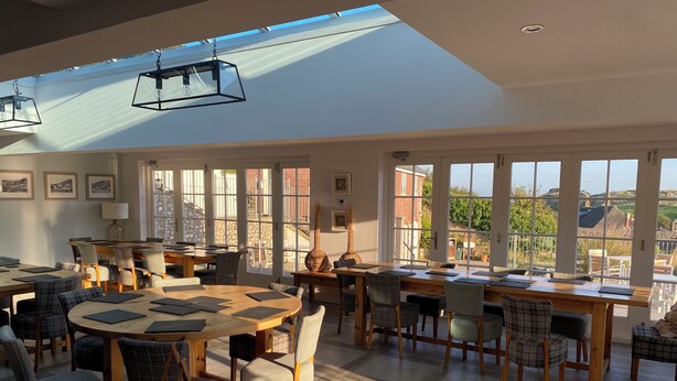 A spacious dining area with multiple tables and chairs, large windows offering views of the countryside, and a skylight above. - Durdle Door Hotel (Wareham)