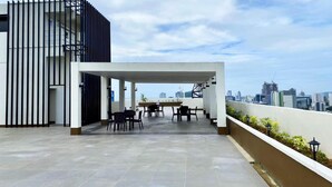 A rooftop terrace with a city view, featuring a covered seating area with tables and chairs.