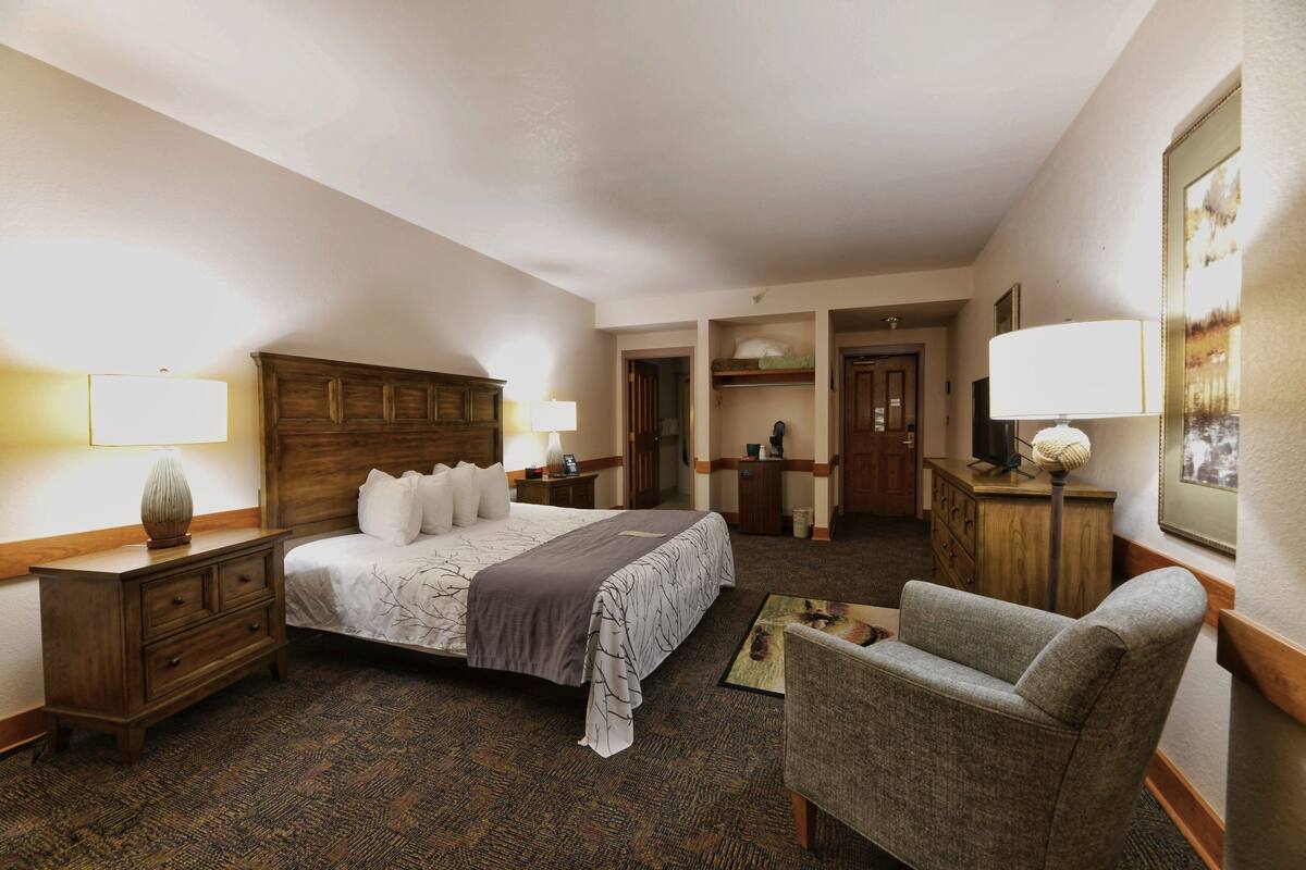 A hotel room with a large bed, a wooden headboard, a desk with a lamp, a chair, and a dresser.